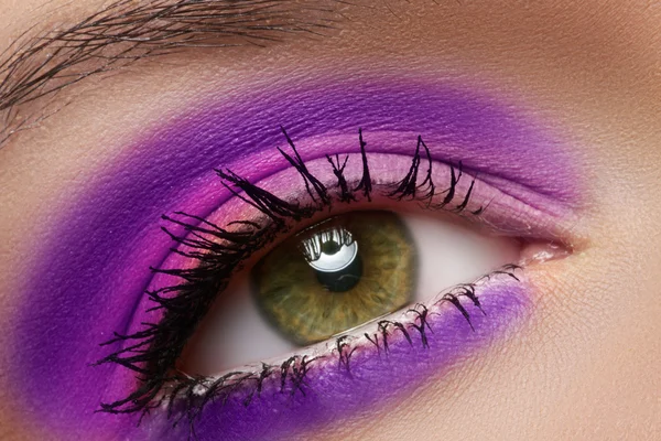 Cosmetics and beauty care. Macro close-up of beautiful green female eye with bright fashion runway make-up. Violet eyeshadows and black eyeliner