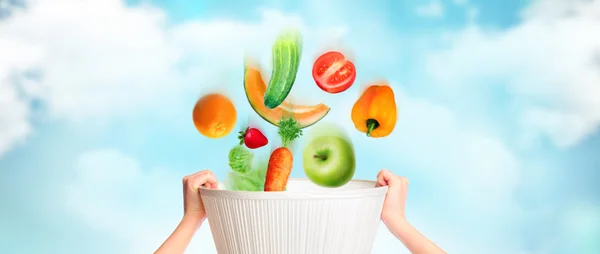 Hands are holding basket against sky background, vegetables, fruits and berries are falling into this basket