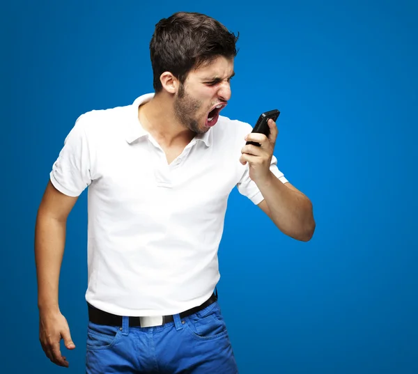Portrait of angry young man shouting using mobile over blue background