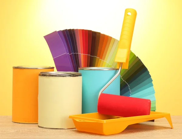 Tin cans with paint, roller, brushes and bright palette of colors on wooden table on yellow background
