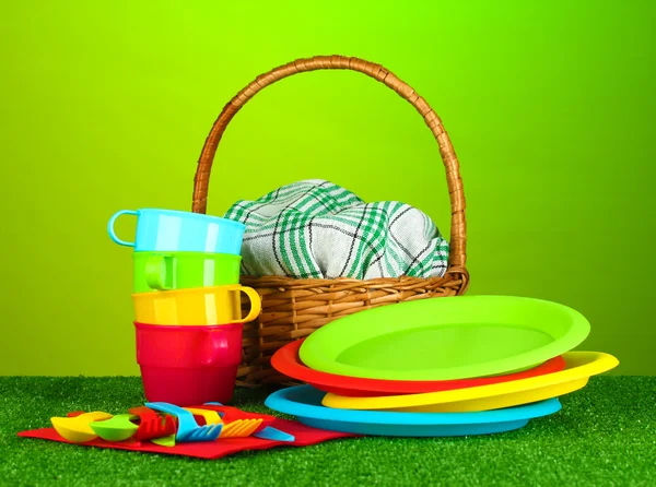 Bright plastic disposable tableware and picnic basket on the lawn on colorful background