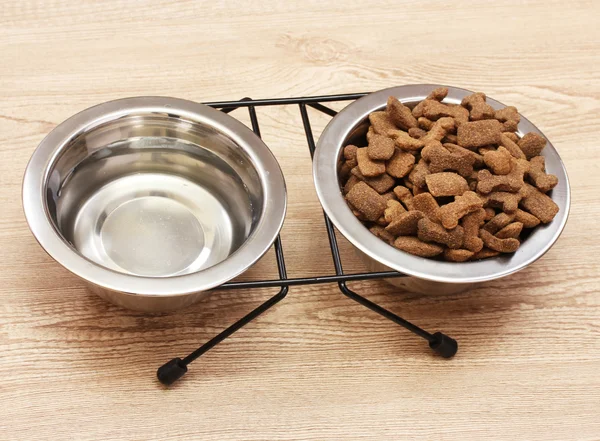 Dry dog food and water in metal bowls on wooden background