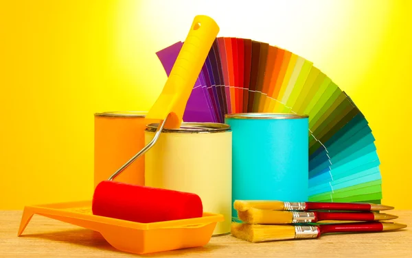 Tin cans with paint, roller, brushes and bright palette of colors on wooden table on yellow background