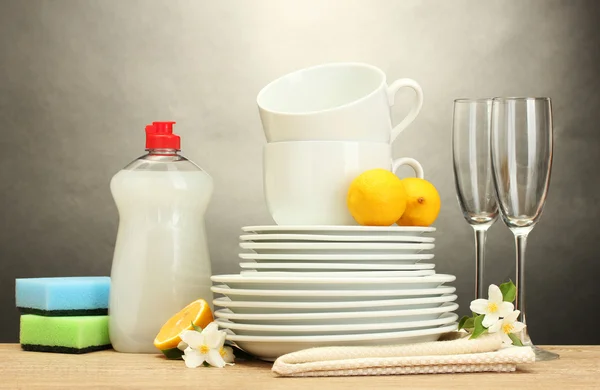 Empty clean plates, glasses and cups with dishwashing liquid, sponges and lemon on wooden table on grey background
