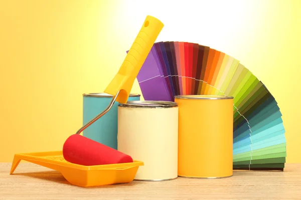 Tin cans with paint, roller, brushes and bright palette of colors on wooden table on yellow background