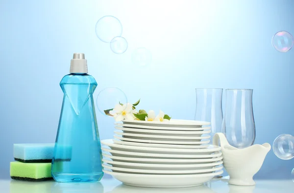 Empty clean plates and glasses with dishwashing liquid, sponges and flowers on blue background