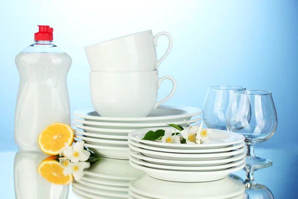 Empty clean plates, glasses and cups with dishwashing liquid and lemon on blue background