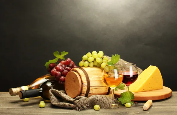 Barrel, bottles and glasses of wine, cheese and ripe grapes on wooden table on grey background