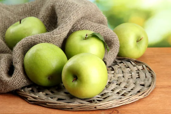 Ripe green apples with leaves on burlap, on wooden table, on green background