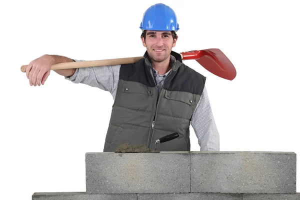 Portrait of handsome bricklayer by brickwall holding shovel over his shoulder