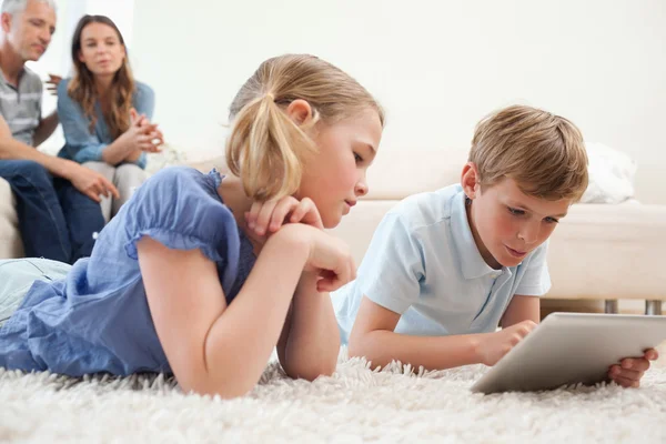 Siblings using a tablet computer with their parents on the backg