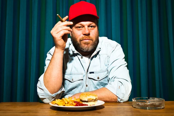 Man with beard eating fast food meal. Enjoying french fries and a hamburger. Smoking a cigar. Trucker with red cap.