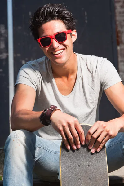 Urban asian man with red sunglasses and skateboard sitting on stairs. Good looking. Cool guy. Wearing grey shirt and jeans.