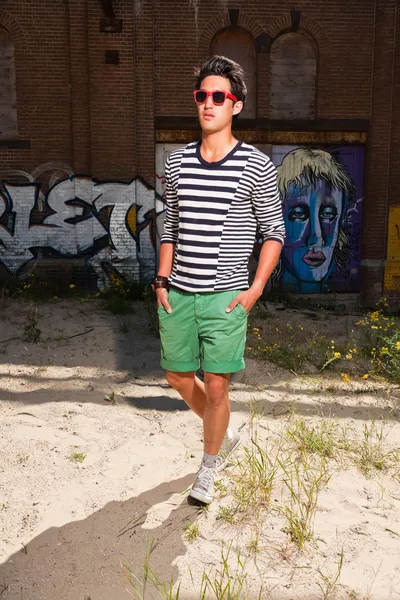 Urban asian man with red sunglasses. Good looking. Cool guy. Wearing blue white striped sweater and green shorts. Standing in front of wall with graffiti.