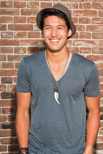 Urban asian man. Good looking. Cool guy. Wearing grey shirt and hat. Standing in front of brick wall.