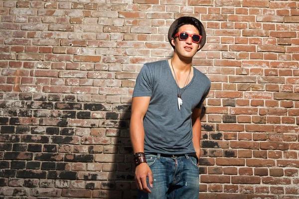 Urban asian man with red sunglasses. Good looking. Cool guy. Wearing grey shirt and hat and jeans. Standing in front of brick wall.