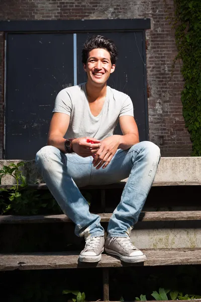 Urban asian man sitting on stairs. Good looking. Cool guy. Wearing grey shirt and jeans. Old neglected building in the background.