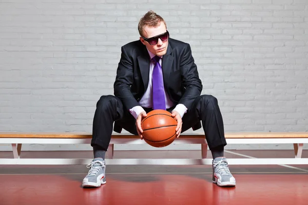 Business man with basketball. Wearing dark sunglasses. Good looking young man with short blond hair. Sitting on bench in gym indoor.