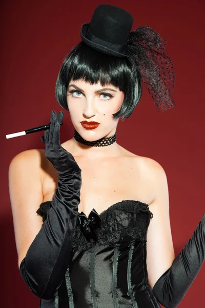 Burlesque pin up woman with black hair dressed in black. Sexy pose. Smoking cigarette. Wearing black hat. Studio fashion shot isolated on red background.
