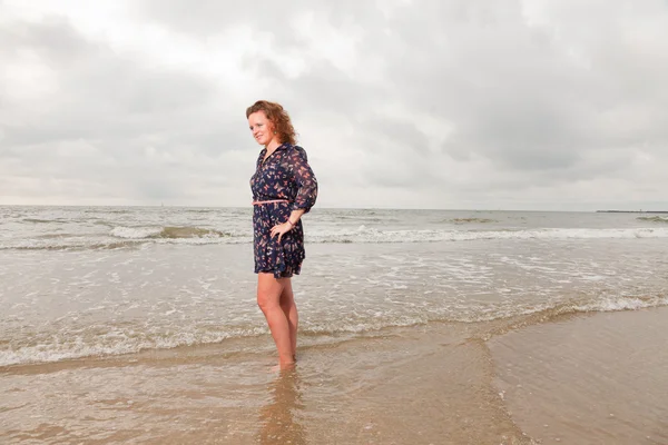 Pretty young woman enjoying outdoor nature near the beach. Standing in the water. Red hair. Wearing dark blue dress. Cloudy sky.