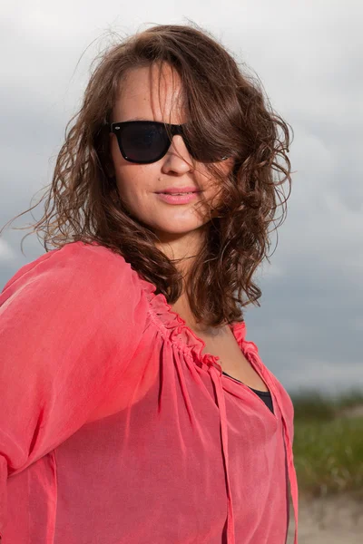 Happy young woman enjoying outdoor nature near the beach. Brown hair. Wearing pink shirt and black sunglasses. Cloudy sky.