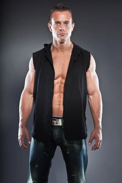 Muscled fitness man. Cool looking. Tough guy. Blue eyes. Blond short hair. Wearing black shirt. Tanned skin. Studio shot isolated on grey background.