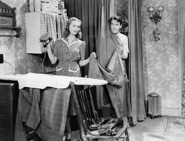 Man and woman standing in a kitchen while she is ironing his pants and he is behind a curtain