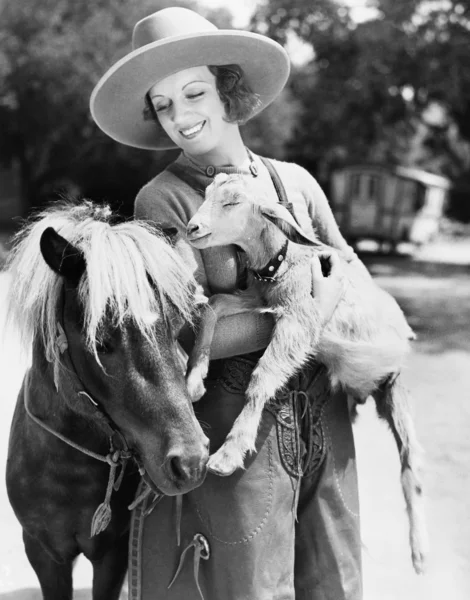 Young woman in a cowboy hat holding a goat while leaning against her pony