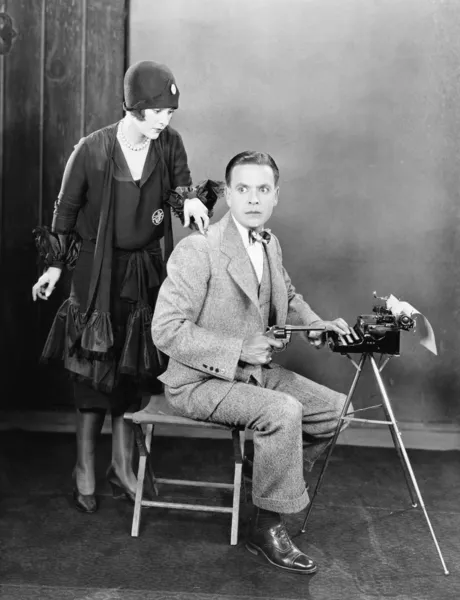 Man at a typewriter holding a gun and a woman tapping on his shoulder