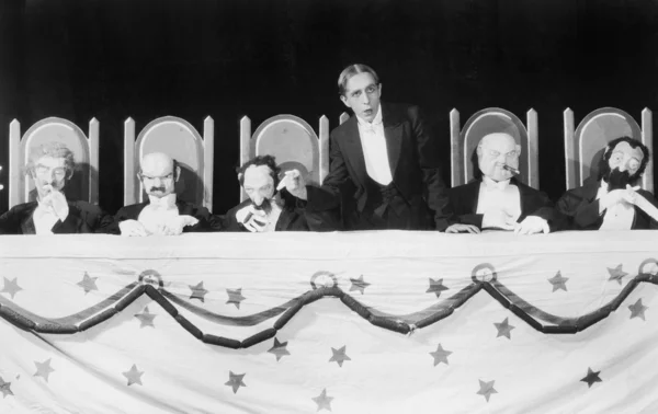Man giving a speech with five character puppets sitting at the table