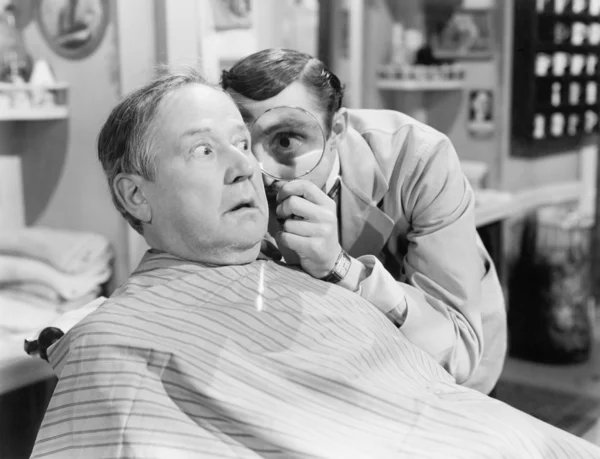 Barber looking at a man's face through a magnifying glass