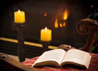 Paperback book open on chair by fire and candle clipart