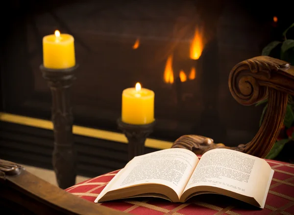 Paperback book open on chair by fire and candle — Stock Photo, Image