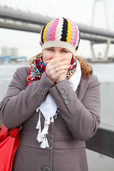Portrait of frozen young Caucasian female warming hands and standing on embankment. Bridge on background — Stock Photo, Image