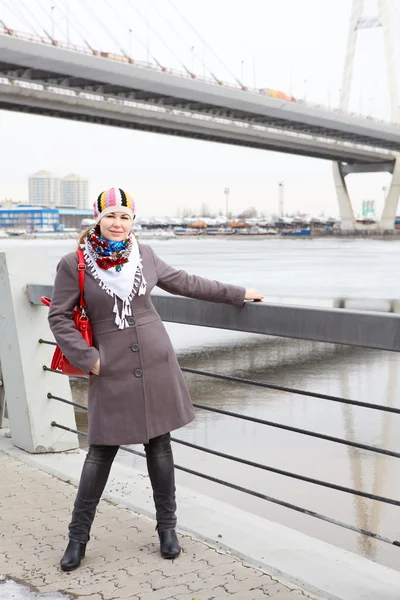 Portrait de heureuse jeune femme caucasienne avec sac rouge debout sur le remblai. Sur toute la longueur. Pont en arrière-plan — Photo