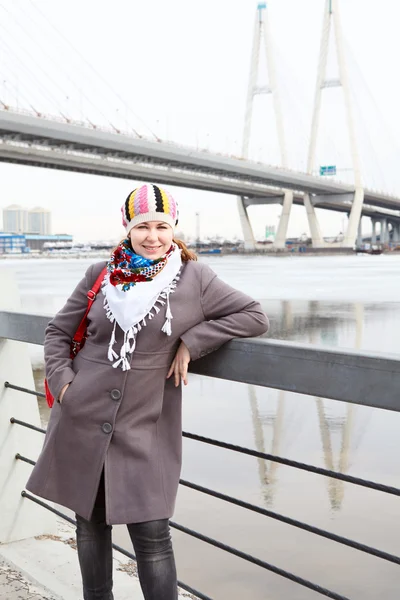 Portrait of happy young Caucasian female with red bag standing on embankment. Bridge on background — Stock Photo, Image