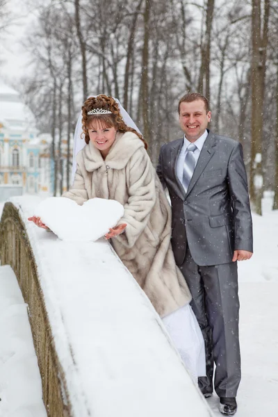 Boda joven pareja rusa caucásica sosteniendo un corazón cubierto de nieve en el puente. Temporada de invierno en Rusia — Foto de Stock