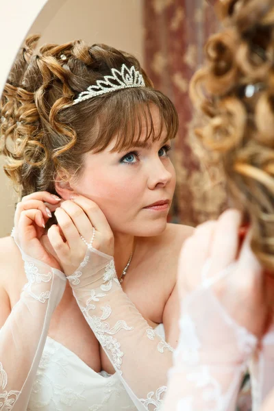 Schöne junge kaukasische Braut mit lockigem Haar, die in den Spiegel schaut und Ohrringe trägt — Stockfoto