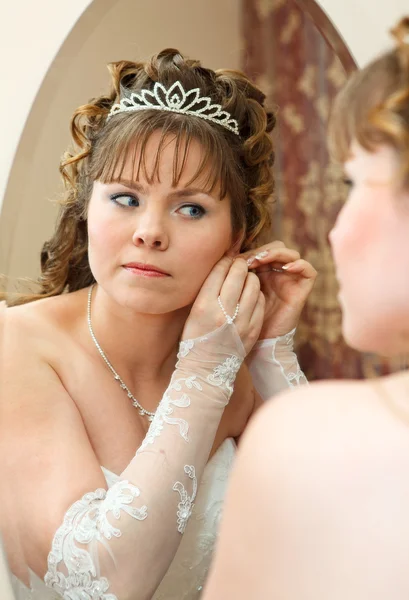 Beleza jovem noiva caucasiana com cabelo encaracolado olhando no espelho e vestindo brincos — Fotografia de Stock