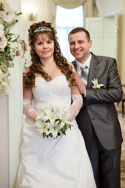 Happy Caucasian newlywed couple standing together. White dress and grey siut. Bunch of lilies in hands — Stock Photo, Image