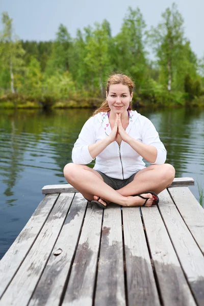 Gelukkige vrouw in lotos pose zittend op houten planken aan rand van de rivier. verticale copyspace — Stockfoto