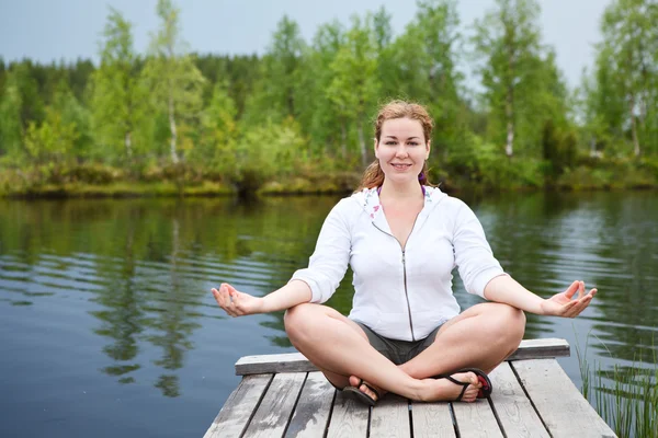 stock image Happy woman in lotos pose sitting on wooden boards on lake edge. Copyspace