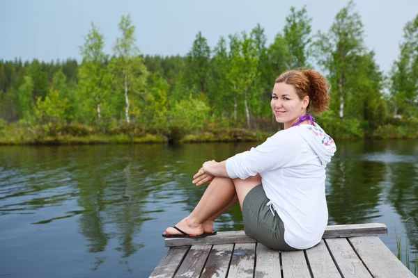 Jovem mulher sorridente sentada em tábuas de madeira na borda do lago — Fotografia de Stock