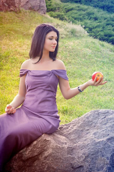 Ung kvinna i klänning sitter på en sten och innehar ett äpple i hans — Stockfoto