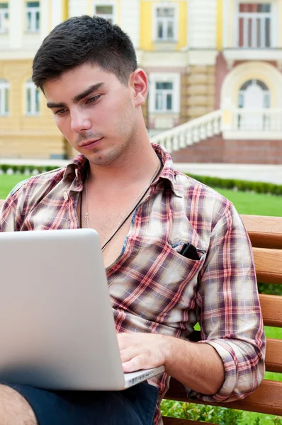 Młody człowiek pracuje na laptopie siedząc na ławce. — Zdjęcie stockowe