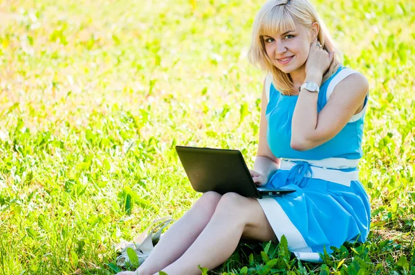 Mulher jovem bonita senta-se em uma grama em um parque com o computador portátil . — Fotografia de Stock