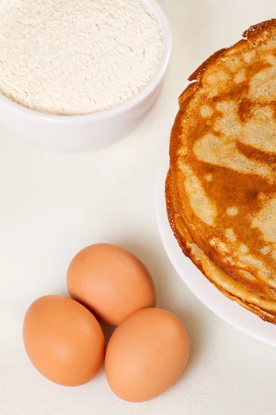 Pile of pancakes with some flour and eggs on white background — Stock Photo, Image