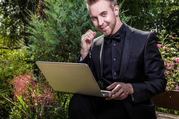 Jonge mooie modieuze man met laptop tegen zomertuin. — Stockfoto