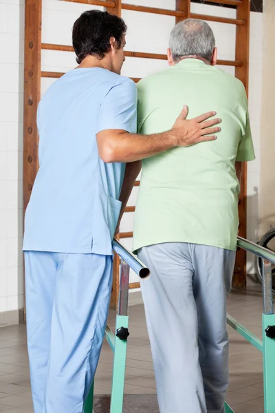 Terapeuta que ayuda al hombre mayor a caminar con el apoyo de las barras — Foto de Stock