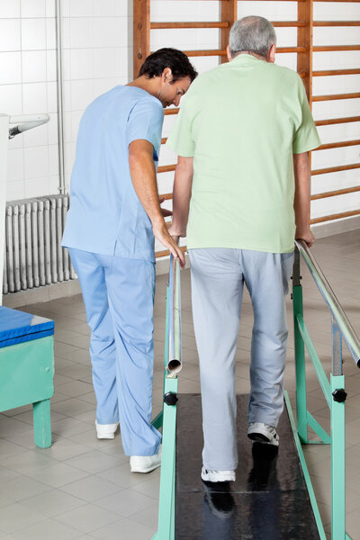 Male Therapist Assisting Senior Man To Walk With The Support Of